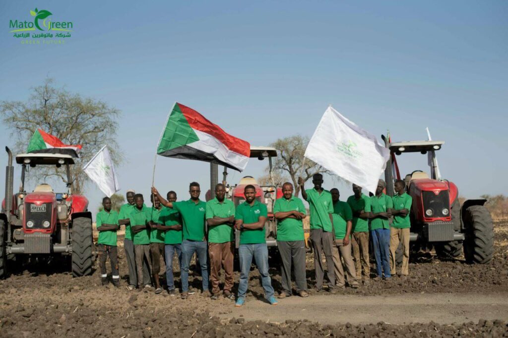مجموعة من الشباب يلبسون ملابس شركة ماتوقرين الخضراء و يرفعون علم شركة ماتوقرين وعلم السودان يقفون على أرض زراعية وخلفهم جرارات وبعض الأشجار